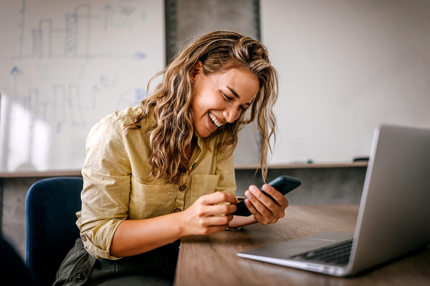 Frau sitzt  lächelnd am Handy, vor ihr steht ein geöffneter Laptop auf dem Tisch